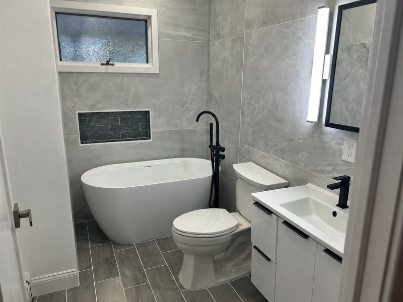 Freestanding soaking tub with black floor-mounted filler, large-format marble-look tile and slate accent niche &mdash; Huntington, NY