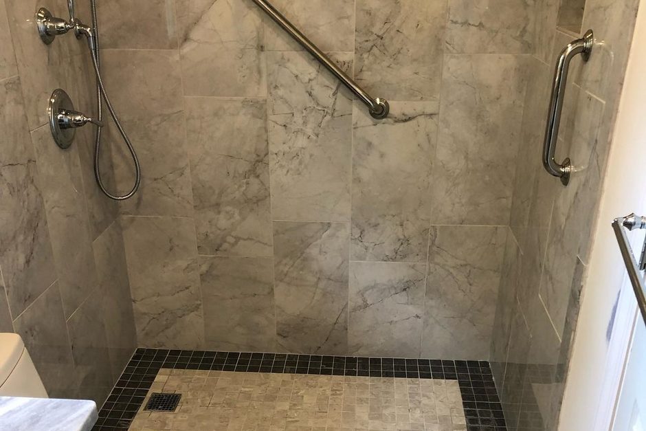 Walk-in tiled shower with two stainless grab bars, a handheld shower on a slide bar and a low mosaic tile entry, installed by Just Enterprise One Corp. on Long Island.