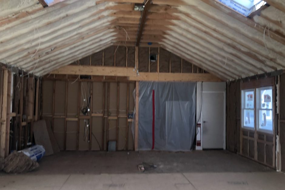 Attached garage-sized space on Long Island mid-conversion into living space: new stud walls, spray foam in the rafters, a plywood subfloor over the slab, a plastic dust barrier at the house door and a new window, by Just Enterprise One Corp.
